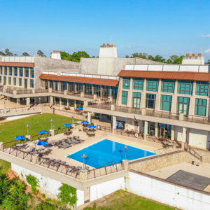 Mountain resort lodge with an outdoor swimming pool. The large building features extensive windows and multiple terraces overlooking a lush green valley. The pool area is surrounded by lounge chairs, tables, and umbrellas, and it sits near the edge of a drop-off, offering dramatic scenic views. Stonework and wooden elements give the lodge a rustic yet modern look, and the grounds are well-maintained with green grass and mature trees. The sky is clear and blue, suggesting a beautiful day.