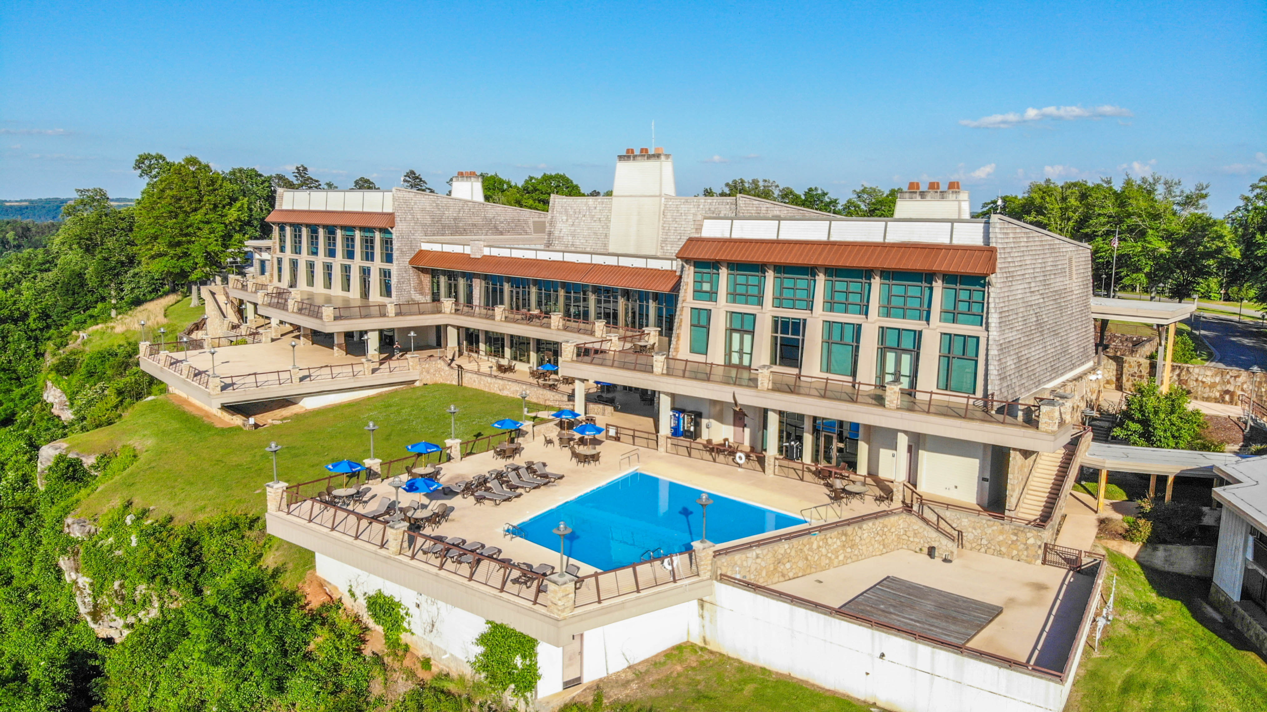 Mountain resort lodge with an outdoor swimming pool. The large building features extensive windows and multiple terraces overlooking a lush green valley. The pool area is surrounded by lounge chairs, tables, and umbrellas, and it sits near the edge of a drop-off, offering dramatic scenic views. Stonework and wooden elements give the lodge a rustic yet modern look, and the grounds are well-maintained with green grass and mature trees. The sky is clear and blue, suggesting a beautiful day.